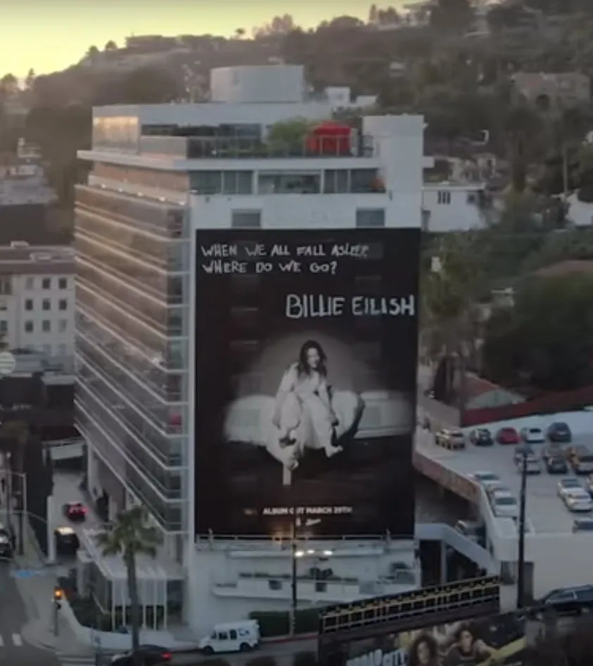 Billboard on a white multi-story building advertising Billie Eilish with the text 'When We All Fall Asleep, Where Do We Go?' in an urban setting.