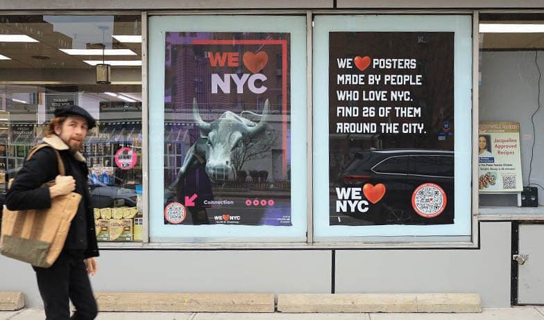 A person carrying a bag walks past a storefront with posters in the windows. One poster features a bull with the text We ❤️ NYC. Another poster says, We ❤️ posters made by people who love NYC. Find 26 of them around the city.