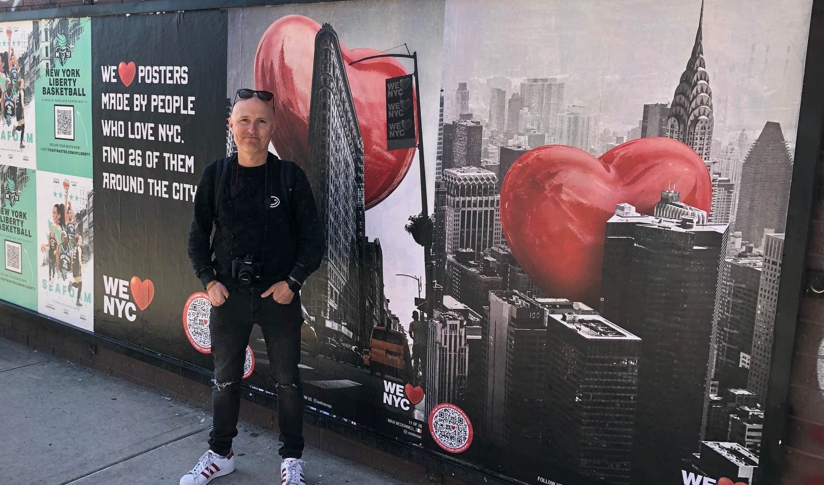 A person stands in front of a wall with NYC-themed posters, featuring red heart motifs integrated into cityscapes, including iconic buildings like the Chrysler Building. The person is wearing sunglasses and casual clothing.