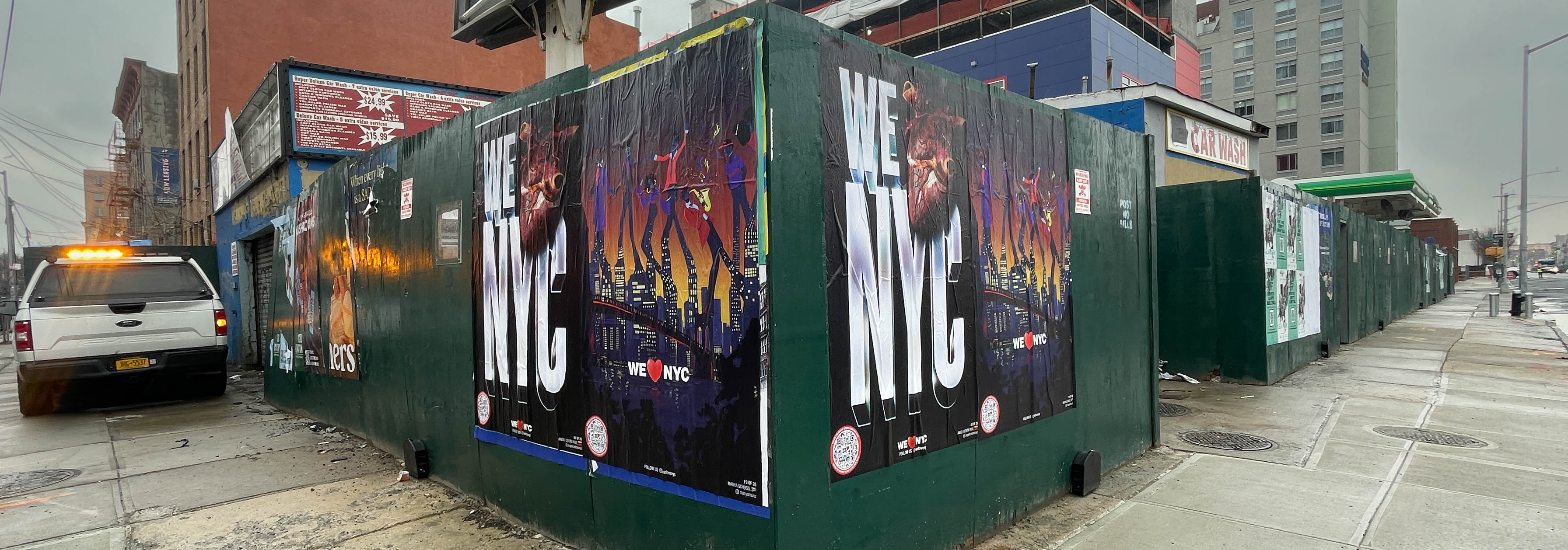 A city street corner with construction barriers covered in posters. The prominent posters display vibrant graphics with the text New NYC. Buildings and a parked vehicle are visible in the background under a cloudy sky.