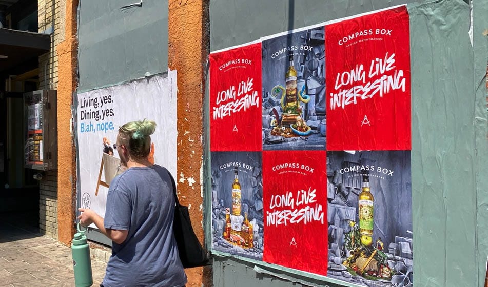 A person with green hair and a blue shirt walks past a wall covered in posters. One poster reads Living, yes. Dining, yes. Blah, nope. Others, labeled COMPASS BOX, feature colorful images and the phrase LONG LIVE INTERESTING.