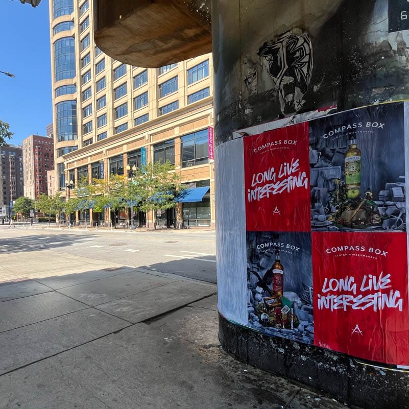 Posters with the text Compass Box: Long Live Interesting are pasted on a concrete pillar beside a sidewalk. A row of trees lines the street, with a multi-story building in the background under a clear blue sky.