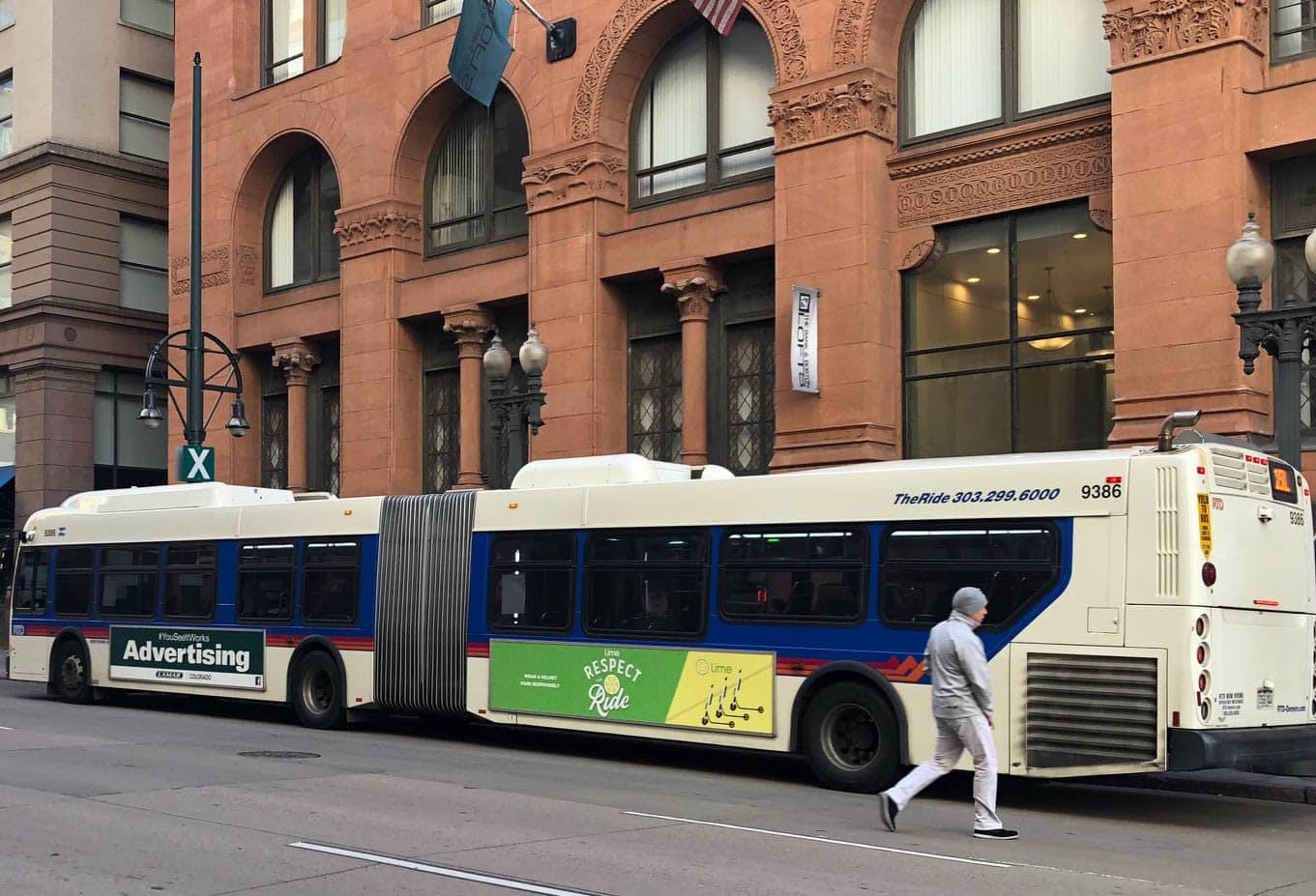 A person in a gray hoodie and white pants walks past an articulated city bus on an urban street. The bus has advertising on its side, and it is parked in front of a building with large arched windows and ornate details.