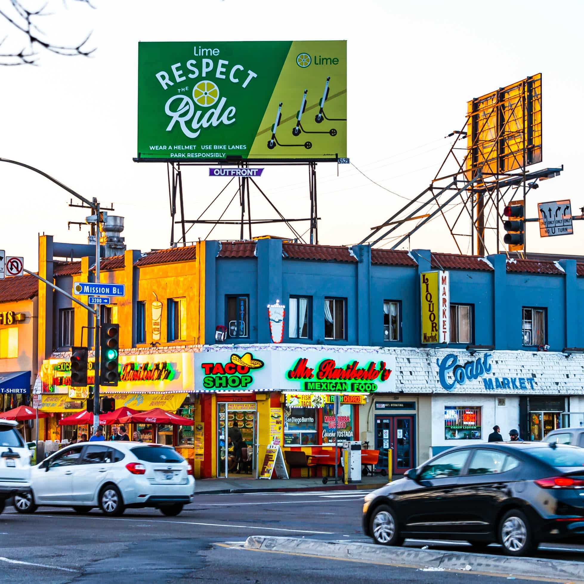 Street scene with a blue building featuring a taco shop and a market on the ground floor. Several cars are on the road in front. A green billboard above promotes Lime scooters, advising to Respect the Ride. Traffic lights and signs are visible.