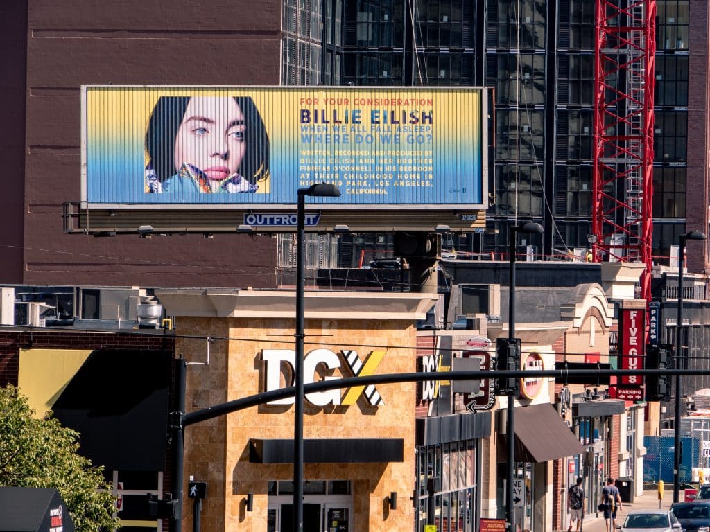 Tri-vision billboard of Billie Eilish promoting her album above a city street with shops and pedestrians in view.