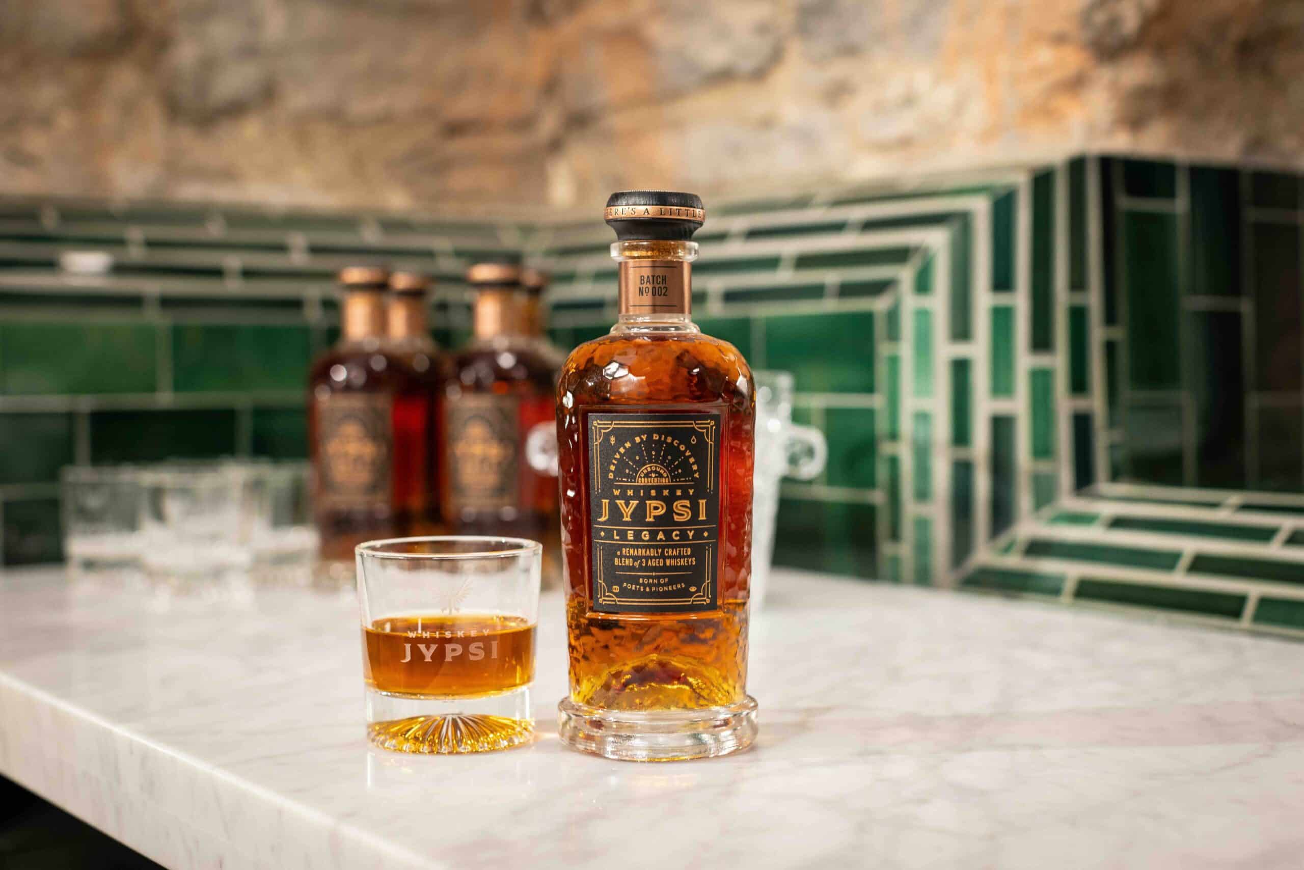 A bottle of JYPSI Legacy rum on a marble countertop, next to a glass filled with rum. In the background, a green tiled wall and more bottles are visible, slightly out of focus.