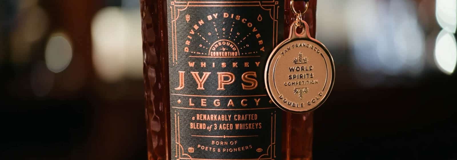 Close-up of a whiskey bottle labeled JYPS Legacy: A Remarkably Crafted Blend of 3 Aged Whiskeys. The bottle features a bronze medal hanging from the neck, awarded for excellence at the World Spirits Competition.