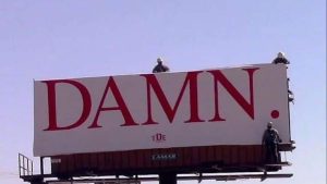 Billboard featuring Kendrick Lamar’s DAMN. album title in red text on white background, with workers installing it in a desert landscape.