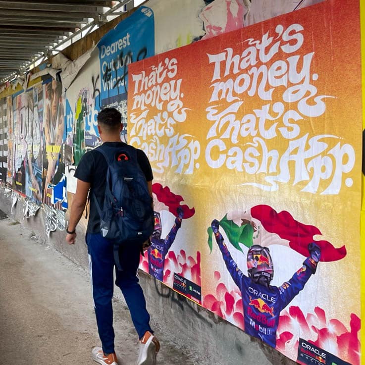A person with a backpack walks past colorful posters on a wall. The posters feature a race car driver celebrating with a flag and the bold text, Thats money. Thats Cash App.
