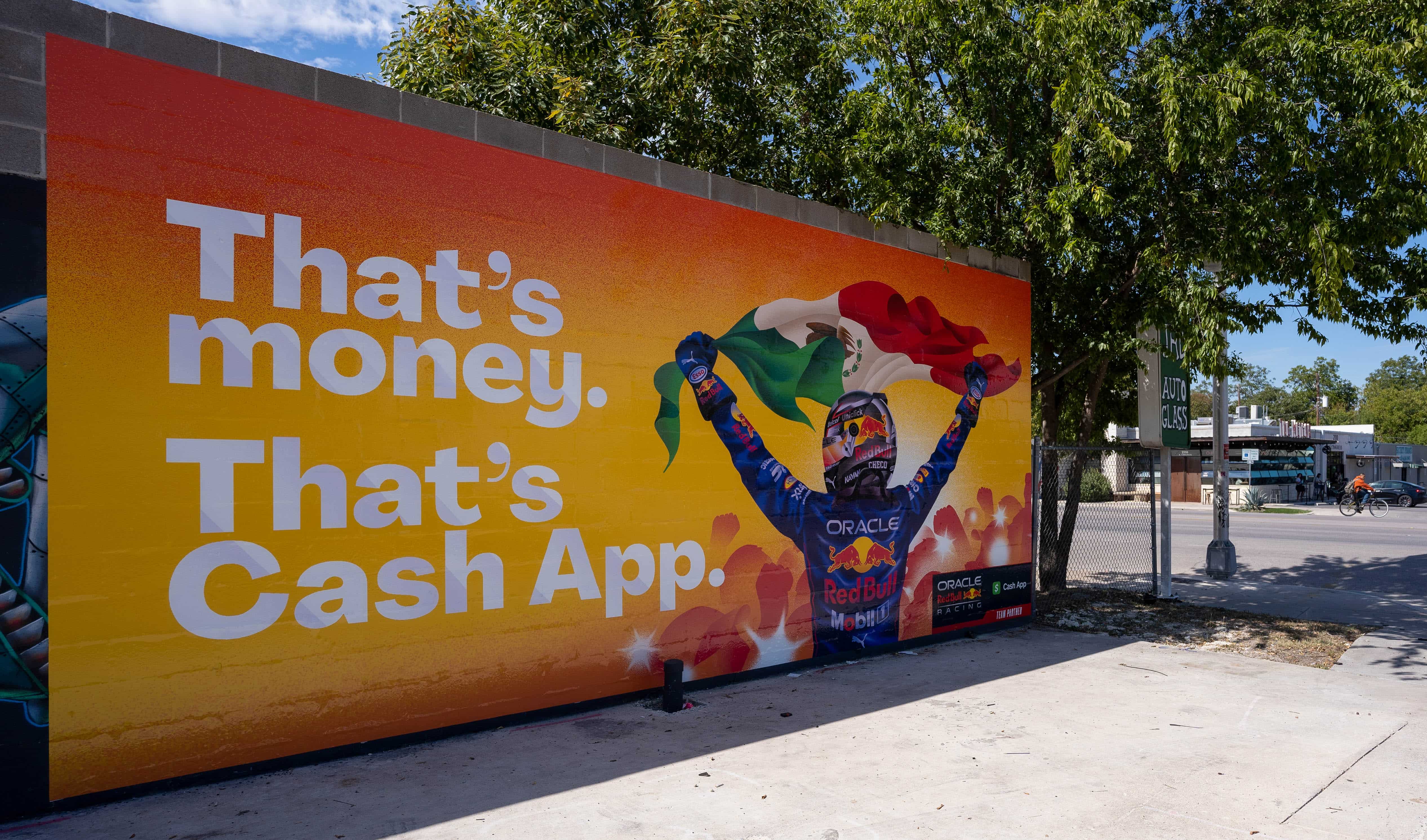 A mural features the text “That’s money. That’s Cash App.” next to an image of a race car driver holding a waving Mexican flag. The mural is outdoors, with trees and a sidewalk nearby.