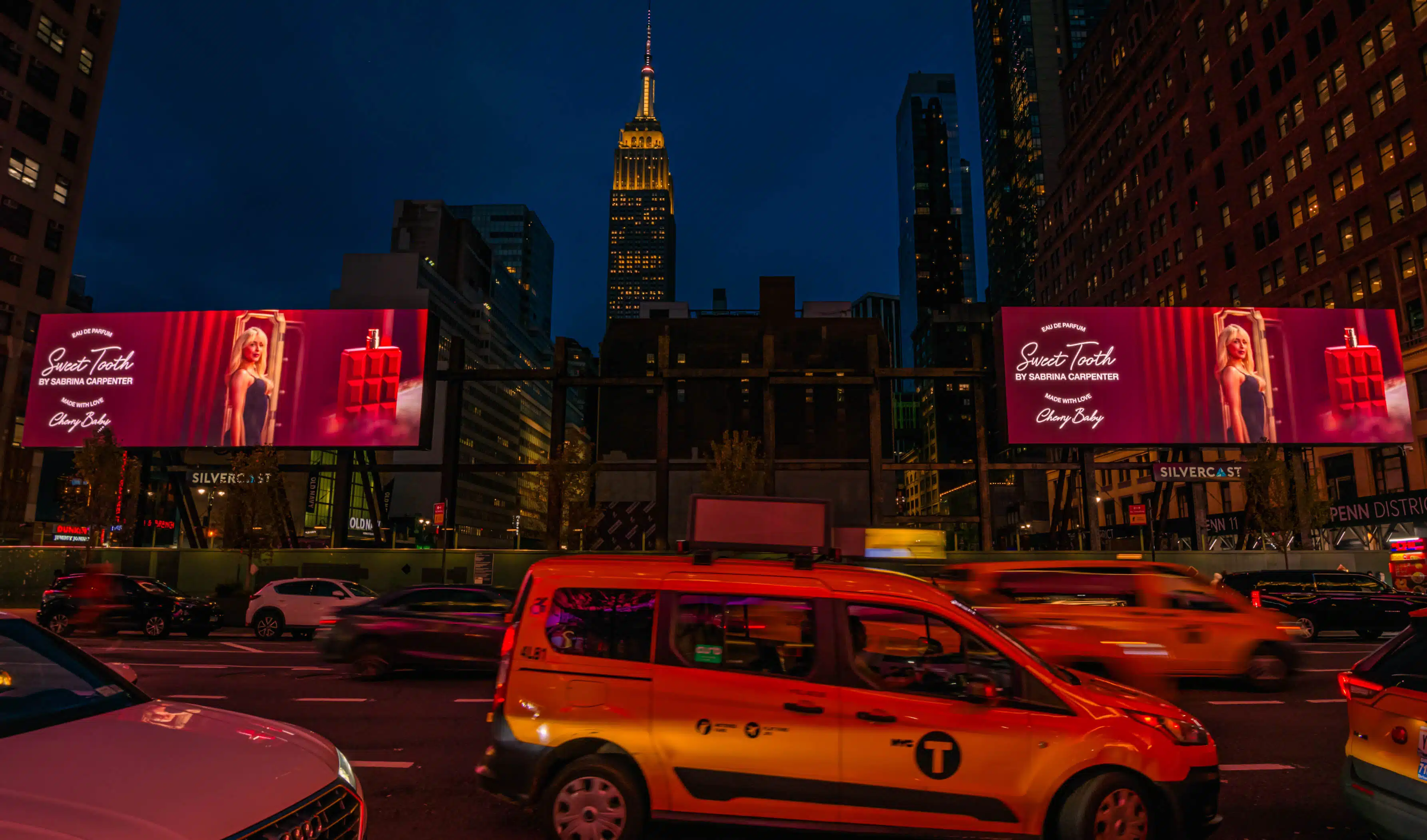 Digital billboard in New York City showing Sabrina Carpenter promoting her Sweet Tooth fragrance, with bright lights and traffic in the foreground.