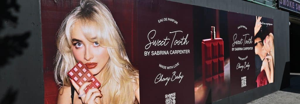 A billboard displays ads for Sweet Tooth by Sabrina Carpenter, featuring a woman holding a chocolate bar and perfume bottles on a red block, with stylized text and QR codes on a dark background.