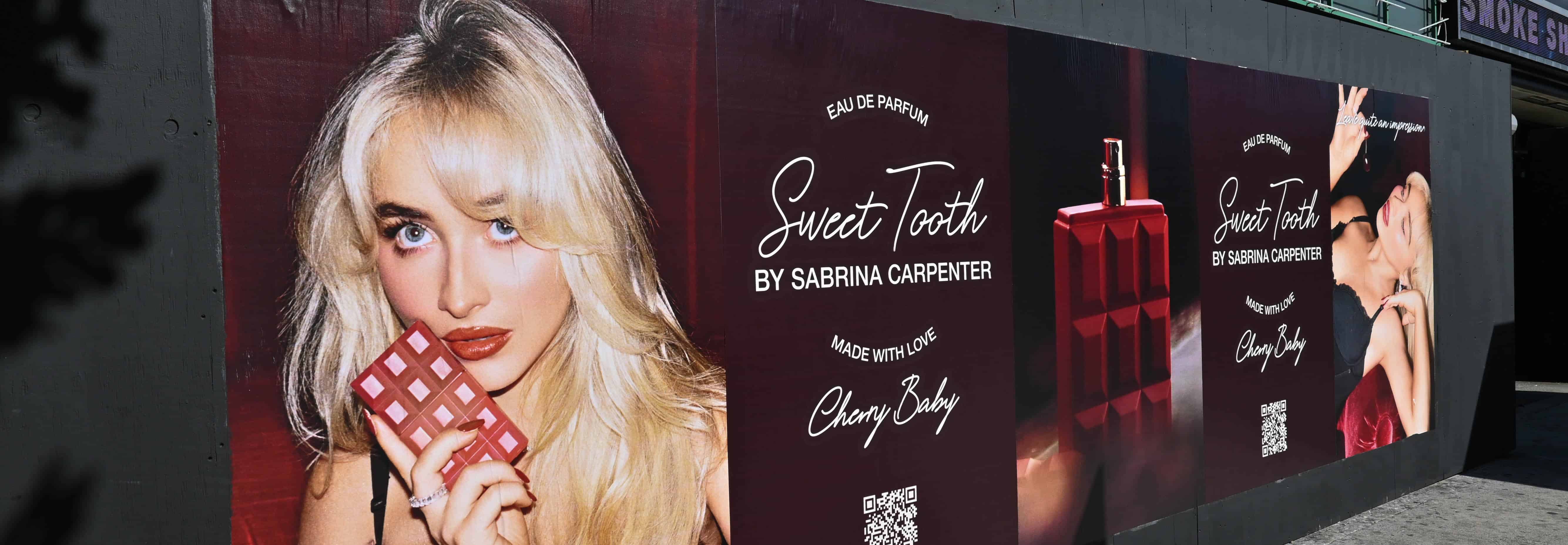 A billboard displays ads for Sweet Tooth by Sabrina Carpenter, featuring a woman holding a chocolate bar and perfume bottles on a red block, with stylized text and QR codes on a dark background.