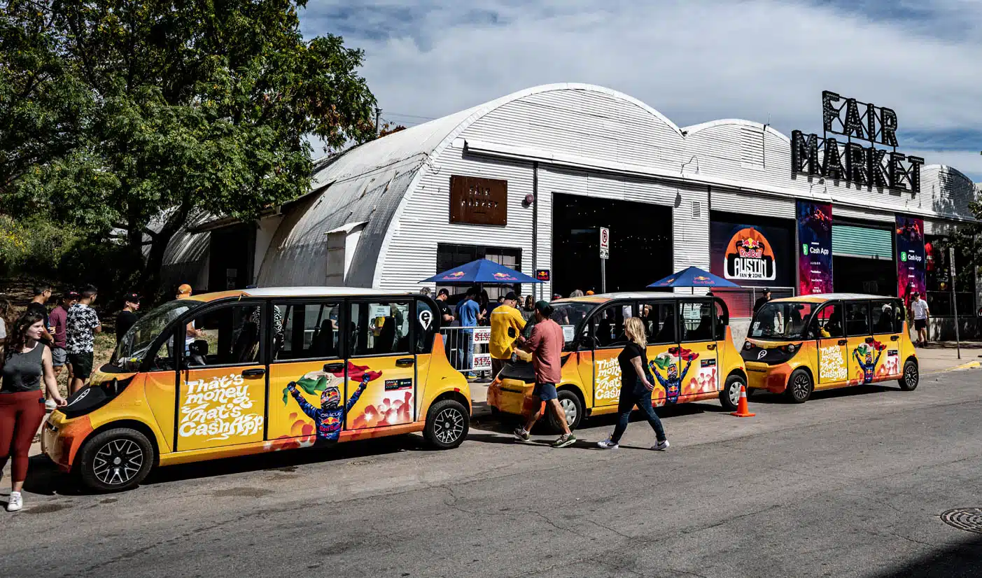 A row of bright yellow electric shuttle vehicles with colorful advertisements parked outside the Fair Market building, as people walk by and gather near the entrance on a sunny day.