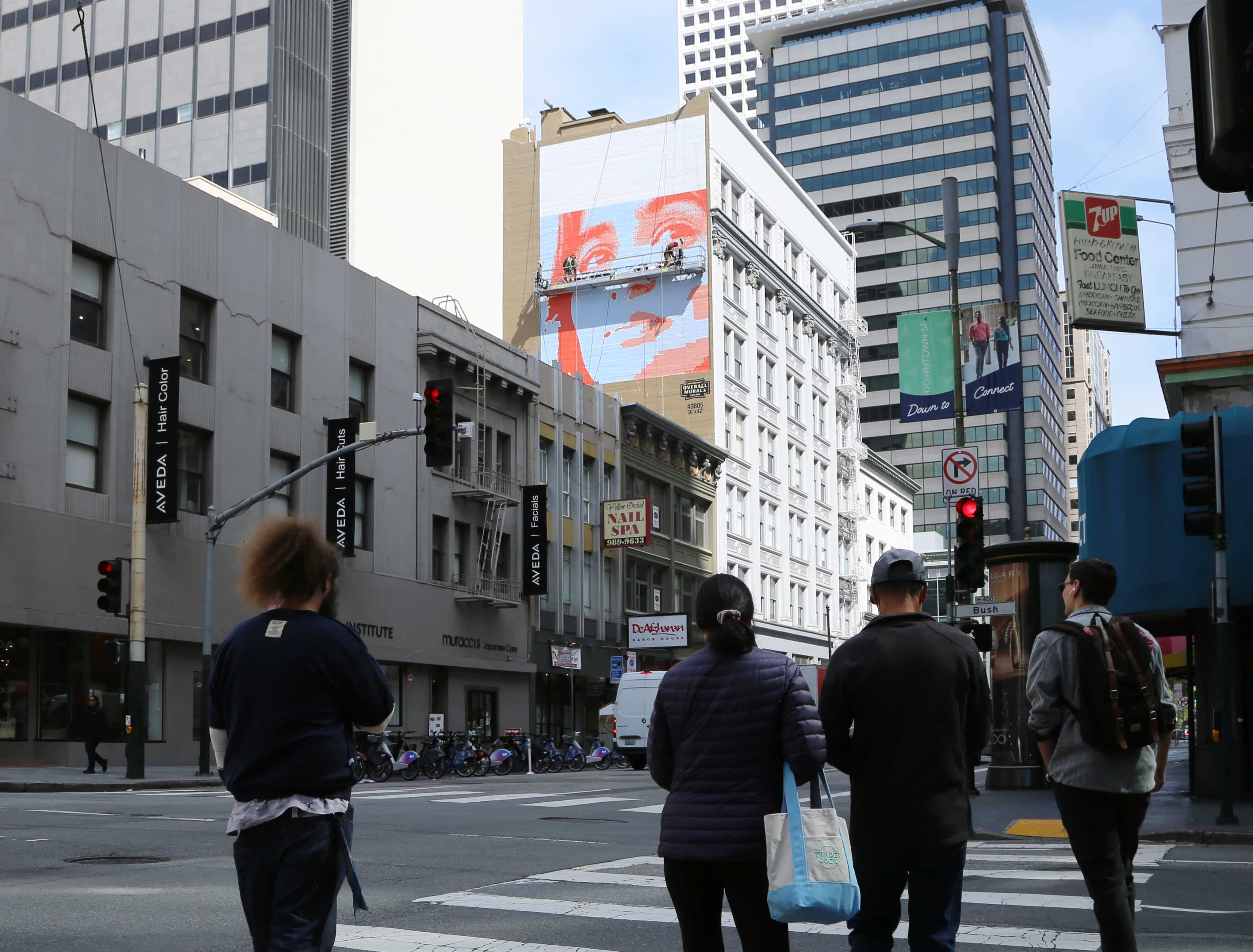 Hand-painted billboard mural in downtown San Francisco, showing large-scale street art installation in progress above pedestrians.