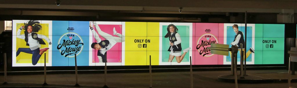 Large digital video wall display in a mall parking garage showing a Disney Club Mickey Mouse campaign