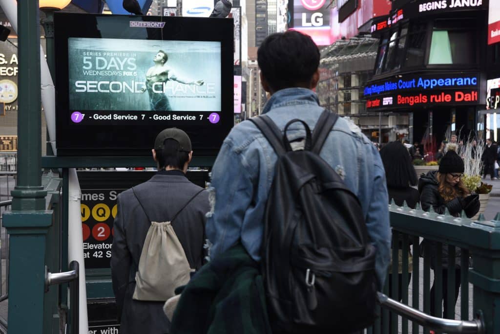 Pedestrians descending into the Times Square–42nd Street subway station in Manhattan, NYC, with an Outfront digital advertising screen displaying a Fox 5 TV show promotion, and illuminated billboards visible in the background.