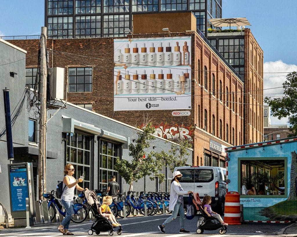 Street-level view of a large ILIA Beauty wall billboard on a brick building in Williamsburg, Brooklyn, displaying foundation serum products with the tagline "Your best skin—bottled," managed by Redrock Outdoor, with street traffic, Citi Bikes, and a pedestrian visible in the foreground.