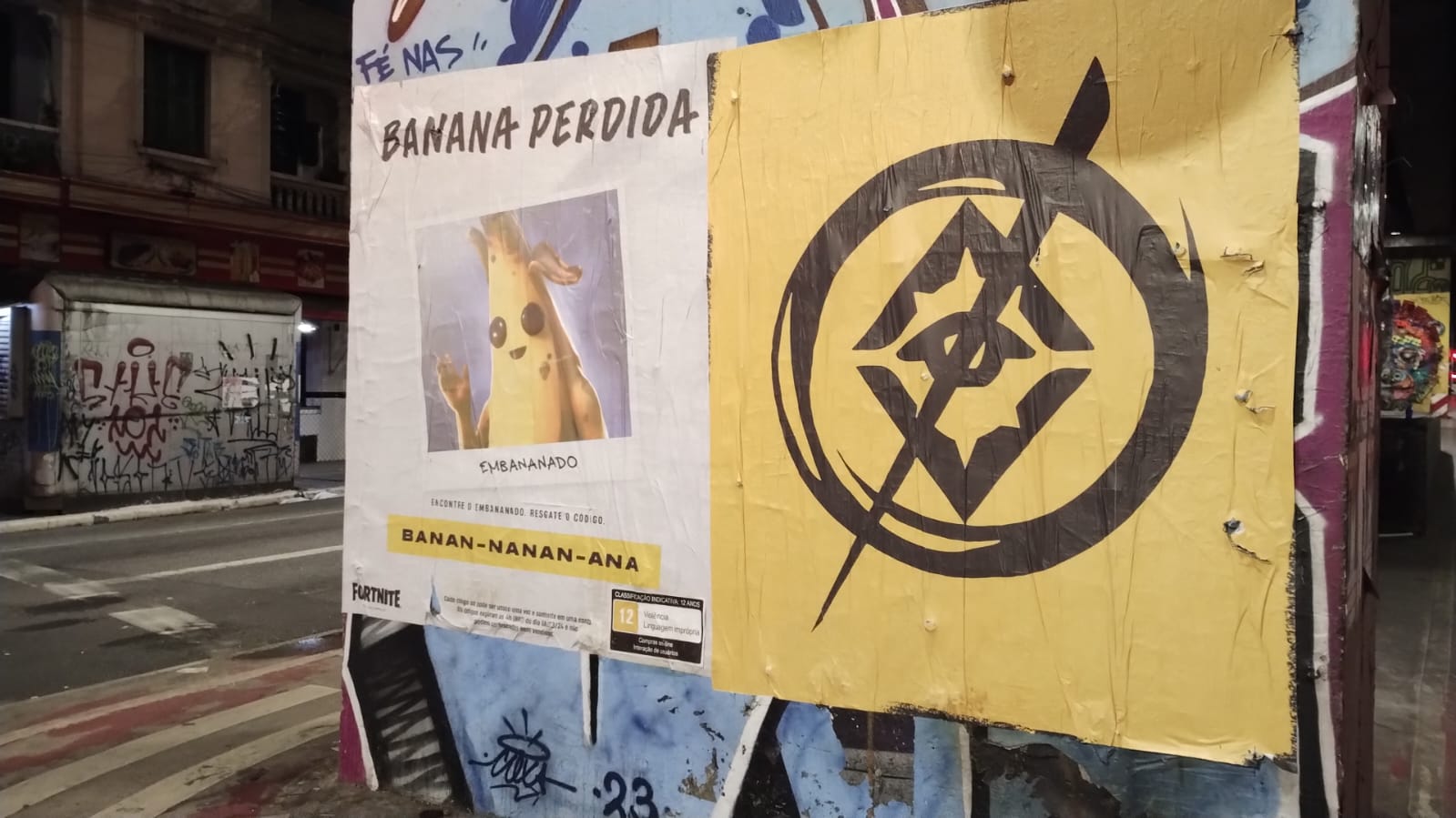 Yellow Fortnite "Banana Perdida" missing poster featuring Peely the banana character next to a stylized eye-and-arrow street art sticker on a graffiti wall in São Paulo, Brazil.