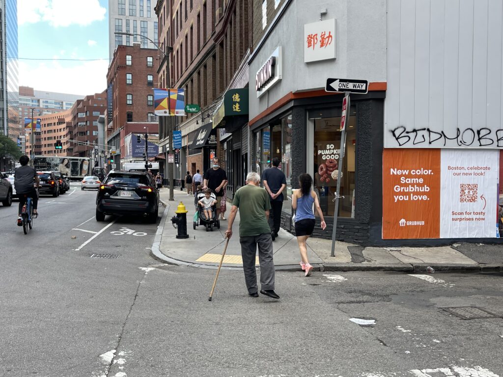 Wild Posting® campaign in Boston's Chinatown at 14 Kneeland Street and Knapp Street — Grubhub "New color. Same Grubhub you love. Boston, celebrate our new look!" rebrand posters with QR code activation, installed on a corner building next to a Dunkin' and steady Chinatown pedestrian foot traffic.
