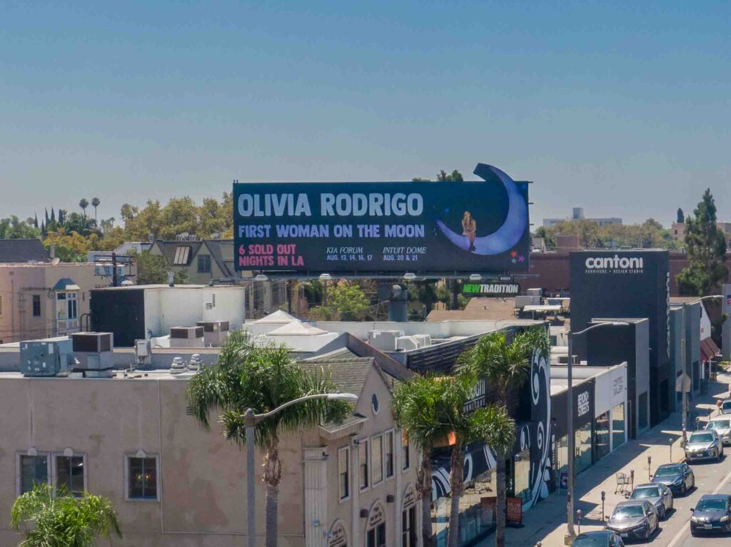 Large billboard reading 'OLIVIA RODRIGO' and 'FIRST WOMAN ON THE MOON' above a city street.
