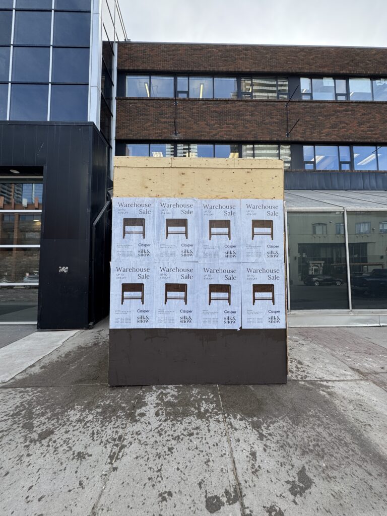Wood-framed storefront under construction with plywood panel and repeated 'Warehouse Sale' posters on the lower panel, urban brick building backdrop.