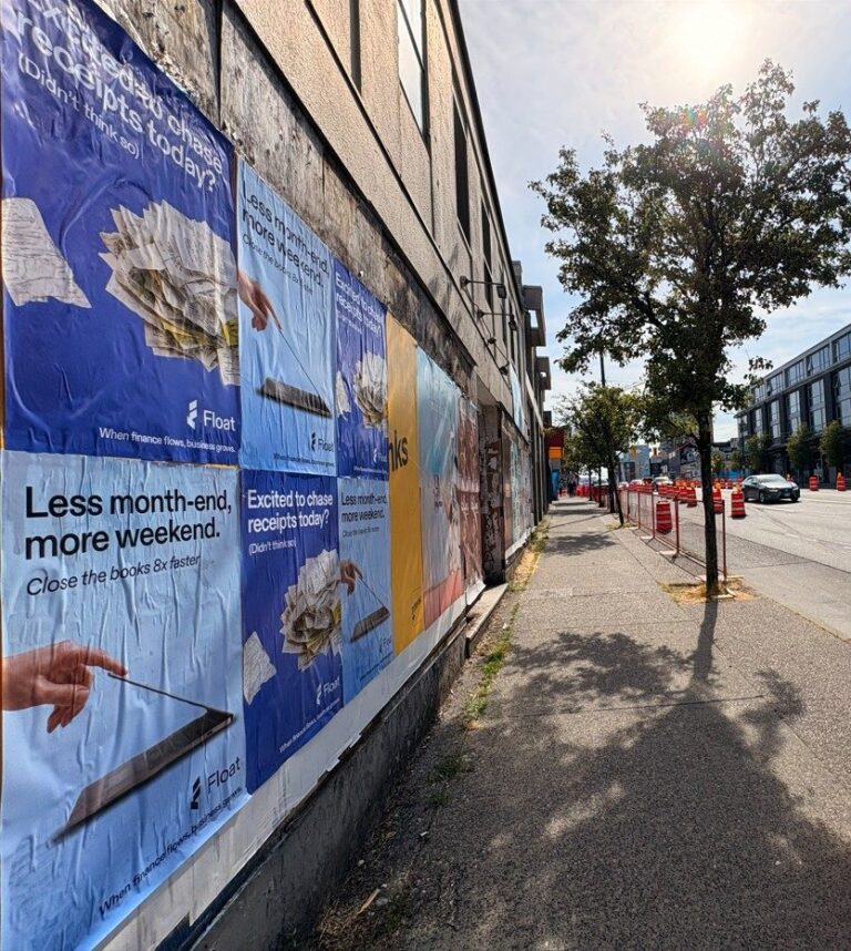Sidewalk along a wall covered in bold blue posters advertising finance offers, with trees and a street in the background.