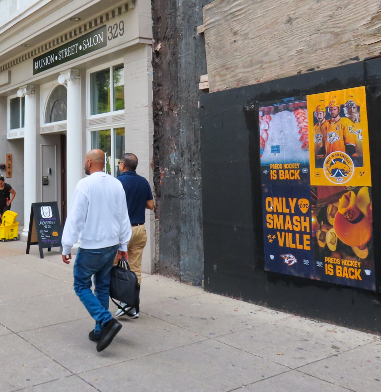 Two men walk past a storefront with a Union Street Salon sign on the left; a construction wall with hockey posters is on the right.