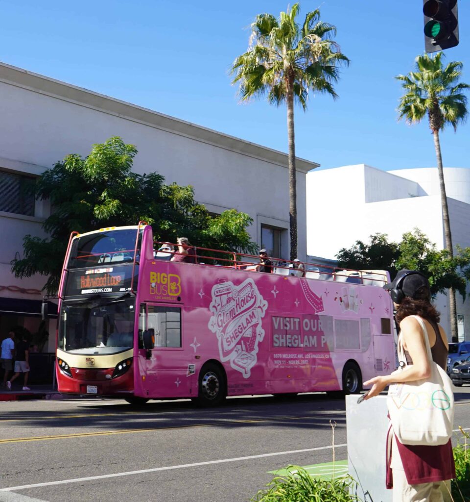 Bright pink double-decker sightseeing bus with graphics driving on a sunny city street, palm trees in the background.