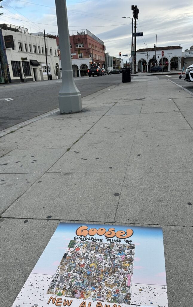 Poster on a sidewalk advertising Goose's new album, with a crowded cartoon collage and a QR code, set against a city street with buildings and a lamppost.