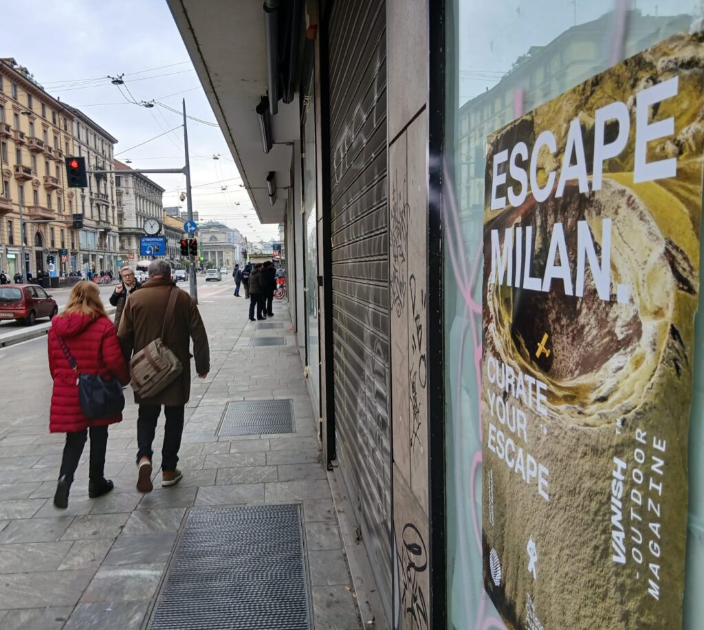 Vanish Outdoor Magazine "Escape Milan — Curate Your Escape" Wheat Pasting and Wild Posting® campaign on a central Milan shopping corridor storefront.
