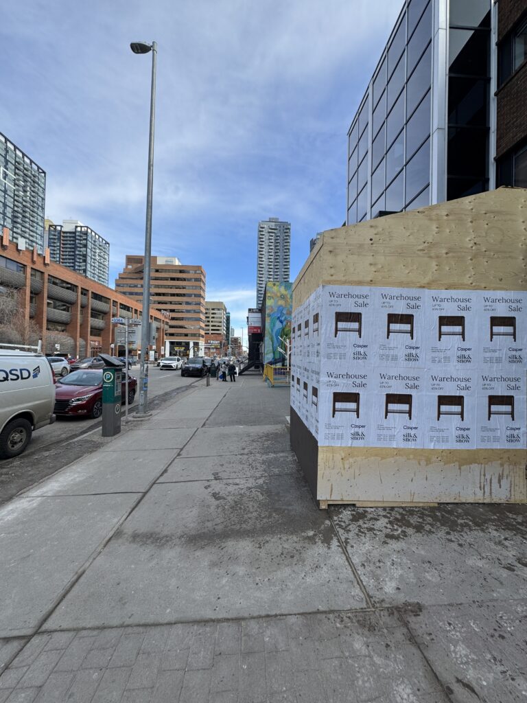 Wild Posting Casper and Silk & Snow Warehouse Sale wheat pasting wrapping a downtown Calgary construction hoarding — eight-poster 4×2 cluster on a corner plywood barricade with downtown Calgary high-rises in the background.