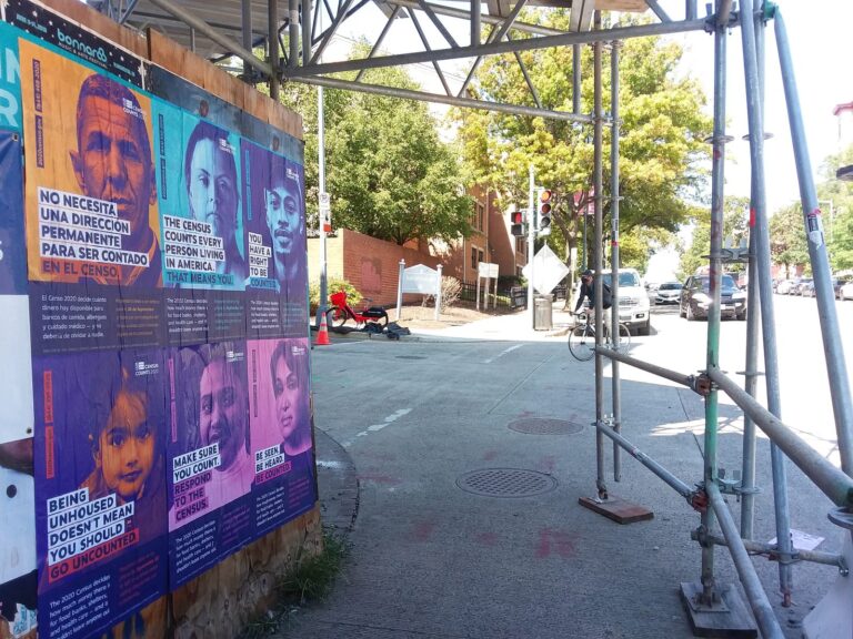 Colorful census posters with portraits on a construction wall along a sidewalk under metal scaffolding at a sunny street corner.