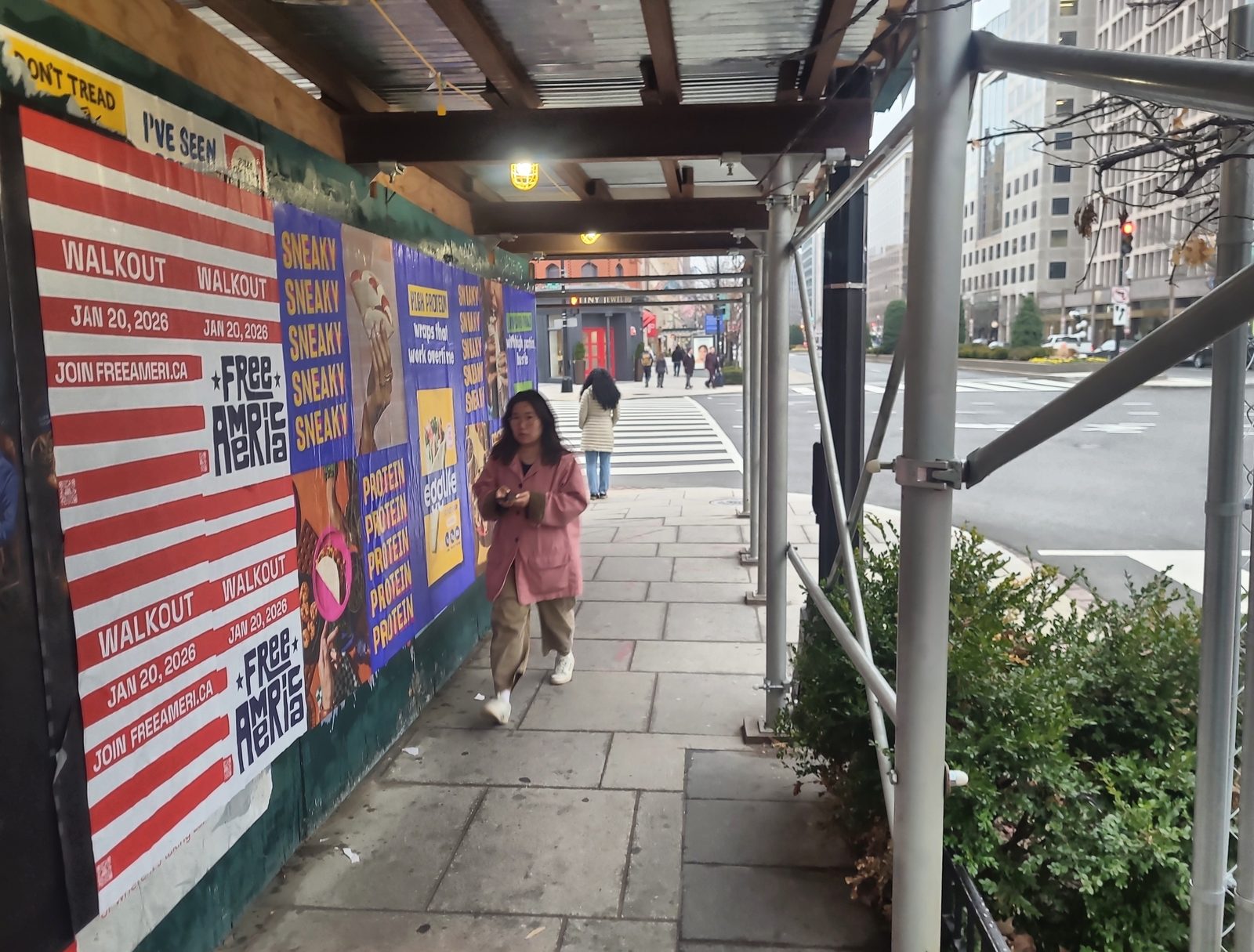 Woman in a pink jacket walks along a sidewalk past a wall covered with colorful posters under scaffolding in a city.