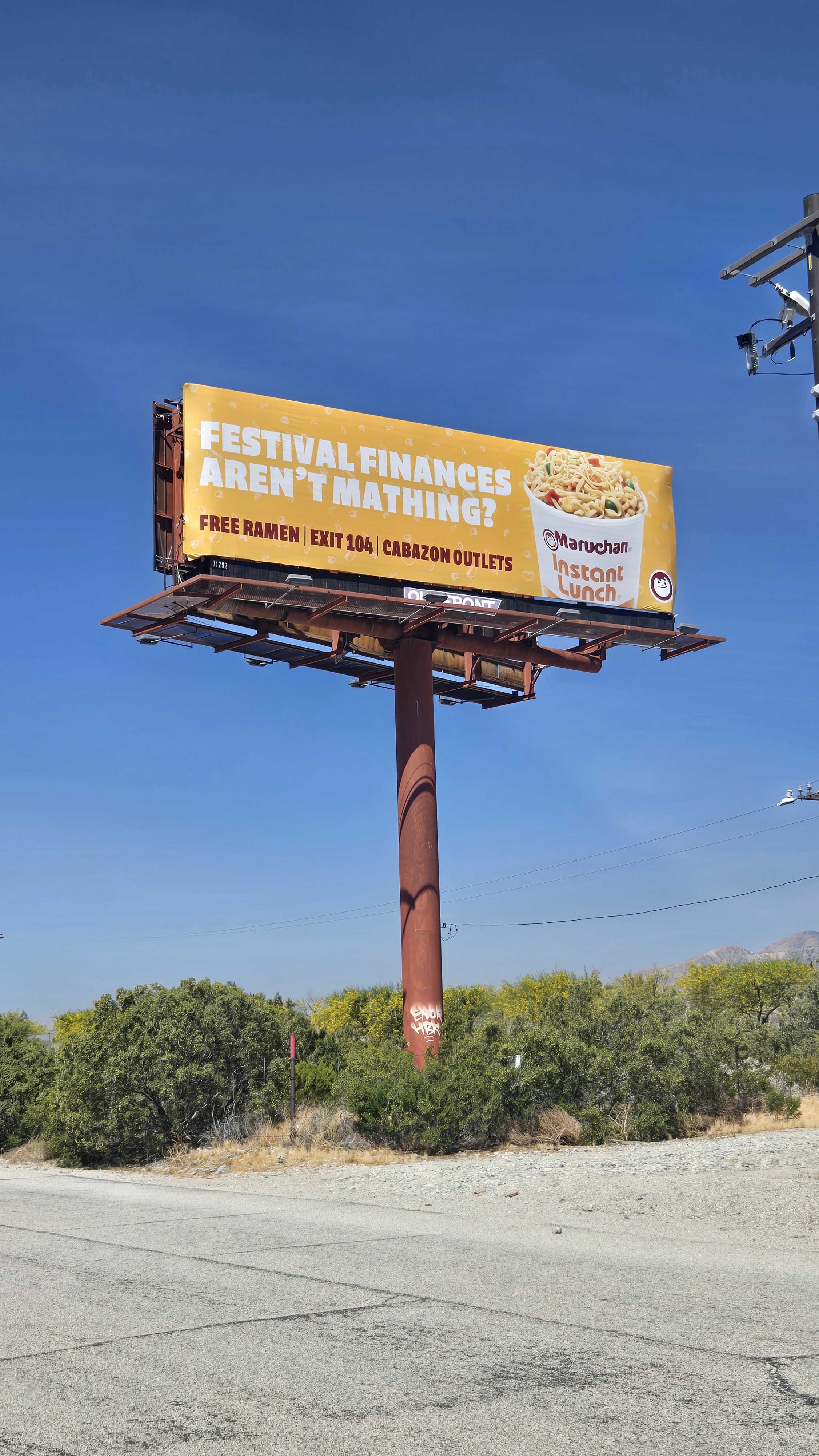 Bright yellow billboard reading 'Festival finances aren't mathing?' with a Maruchan Instant Lunch cup image on the right.