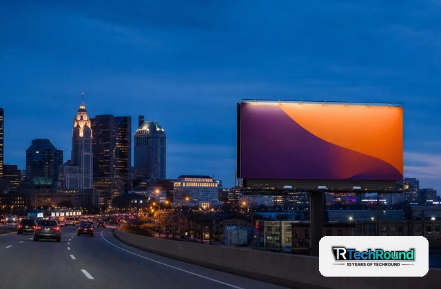 Evening city highway with a large colorful billboard on the right against a blue sky and skyline in the background, watermark in the lower-right corner.