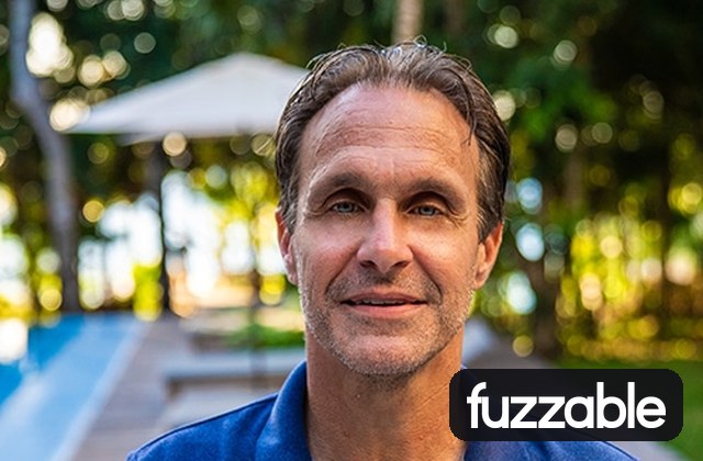 Portrait of a smiling middle-aged man in a blue shirt outdoors by a pool with greenery in the background, and a 'fuzzable' watermark in the corner.