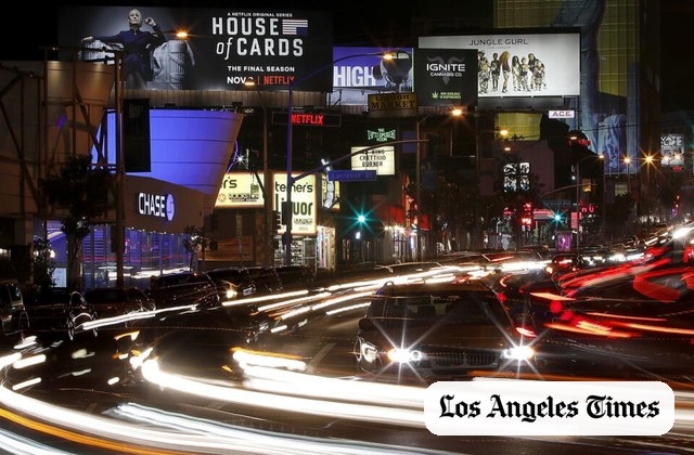 Busy nighttime Los Angeles street scene with bright billboards and light trails from speeding cars; Los Angeles Times logo in the lower-right corner.
