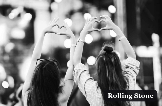 Two friends seen from behind raise their arms to form a heart shape at a lively event, with a Rolling Stone label in the corner.