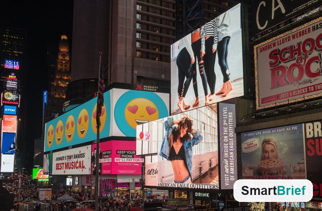 Nighttime view of Times Square with brightly lit billboards, including a row of yellow emoji ads and fashion and Broadway signs, and a crowded pedestrian area in the foreground.