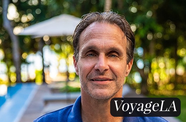 Portrait of a middle-aged man in a blue shirt outdoors, smiling, with a blurred park backdrop and a VoyageLA watermark in the corner.