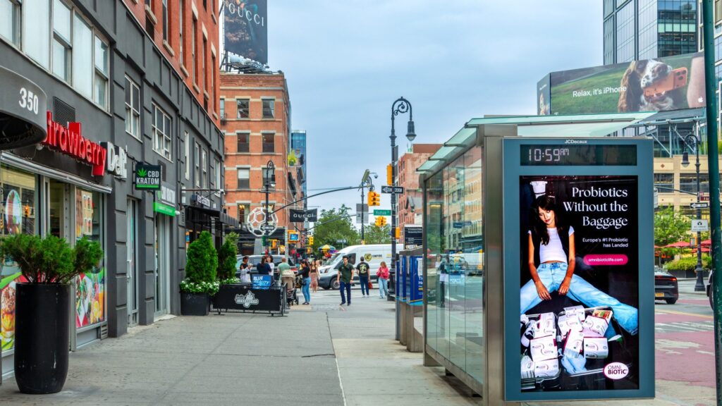 Busy city sidewalk with storefronts, pedestrians, and a glass bus shelter displaying a probiotic ad.