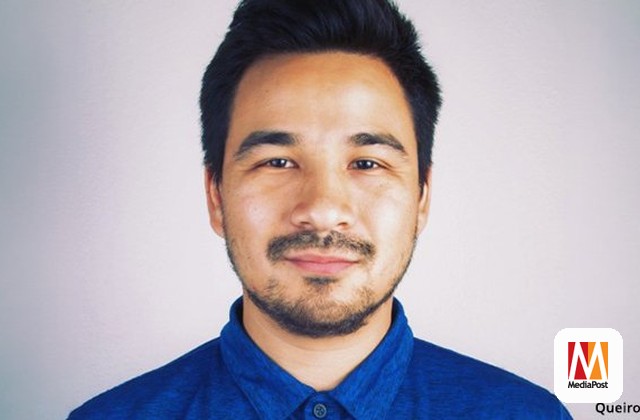 Portrait of a man with short black hair and light stubble, wearing a blue button-down shirt, smiling at the camera against a light gray background.